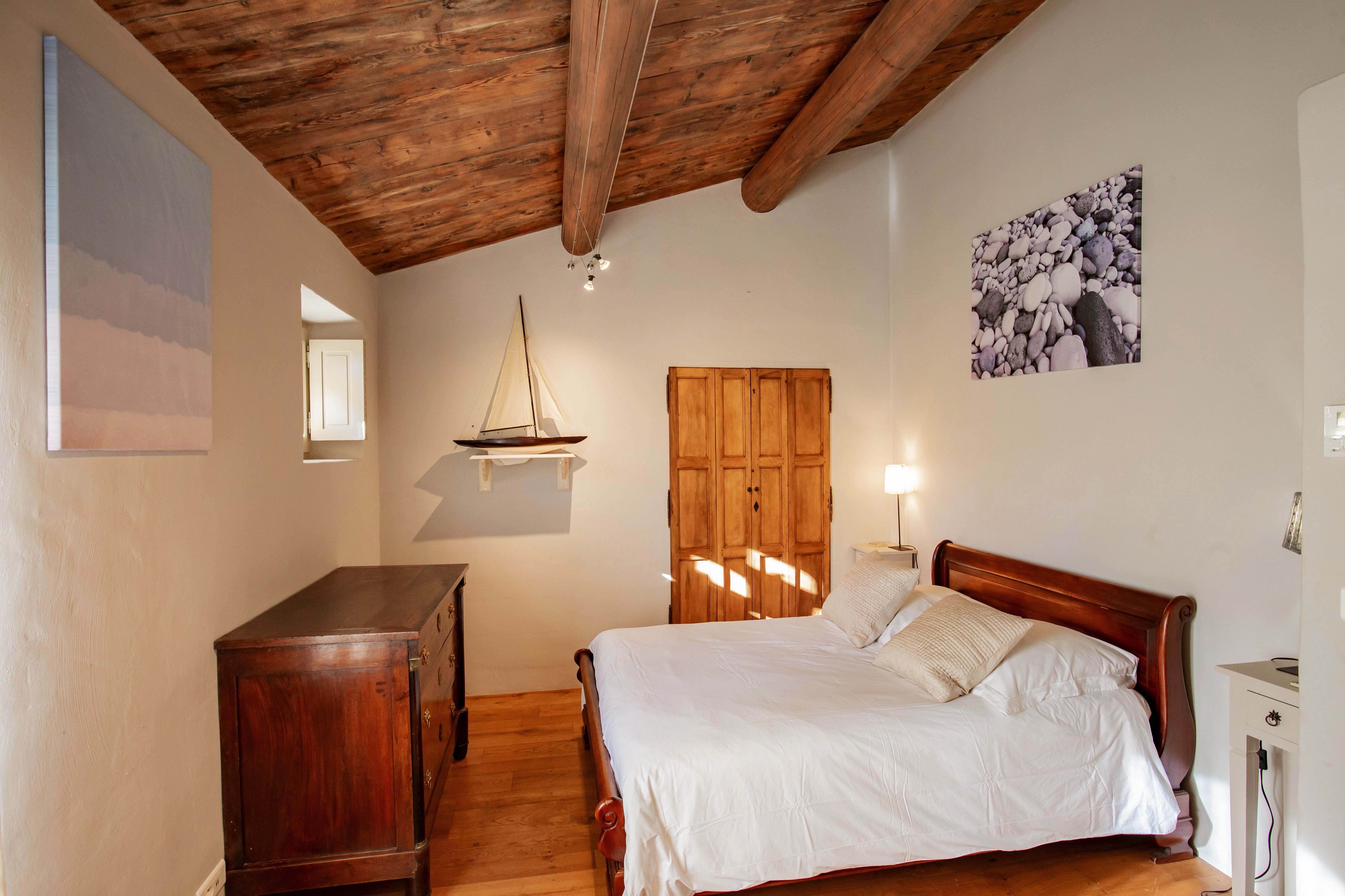 A cozy bedroom featuring a wooden ceiling with beams, a double bed with white bedding, a wood-paneled wardrobe, antique wooden dresser, and wall art including a sailboat and a landscape.